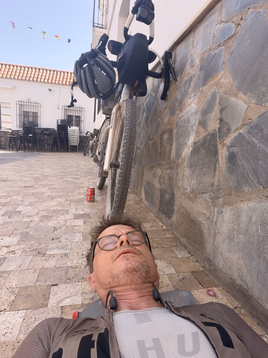 A rider lying on his back on a tiled plaza, the loaded bike leaning on a stone wall above him, a can of cola on the ground.