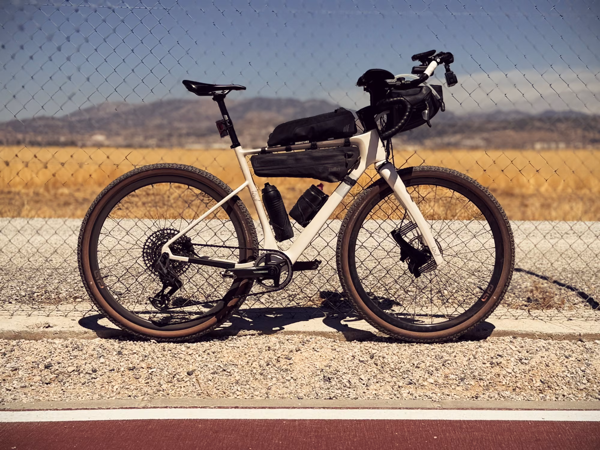 A cream-colored gravel bike with bags and aerobars leans against a chain-link fence, with dry fields and mountains beyond.