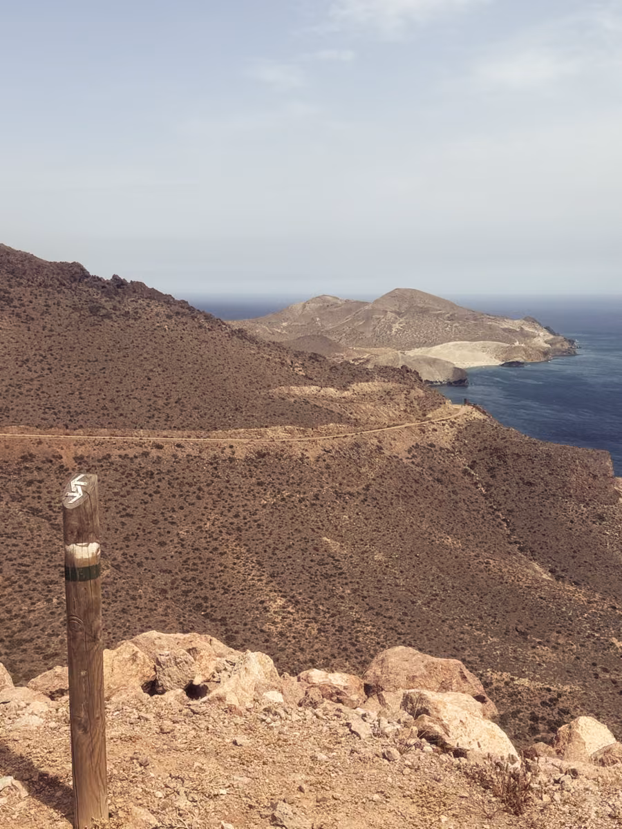 A dirt coast road along brown hills above a blue sea, a headland in the distance, a trail marker post in the foreground.