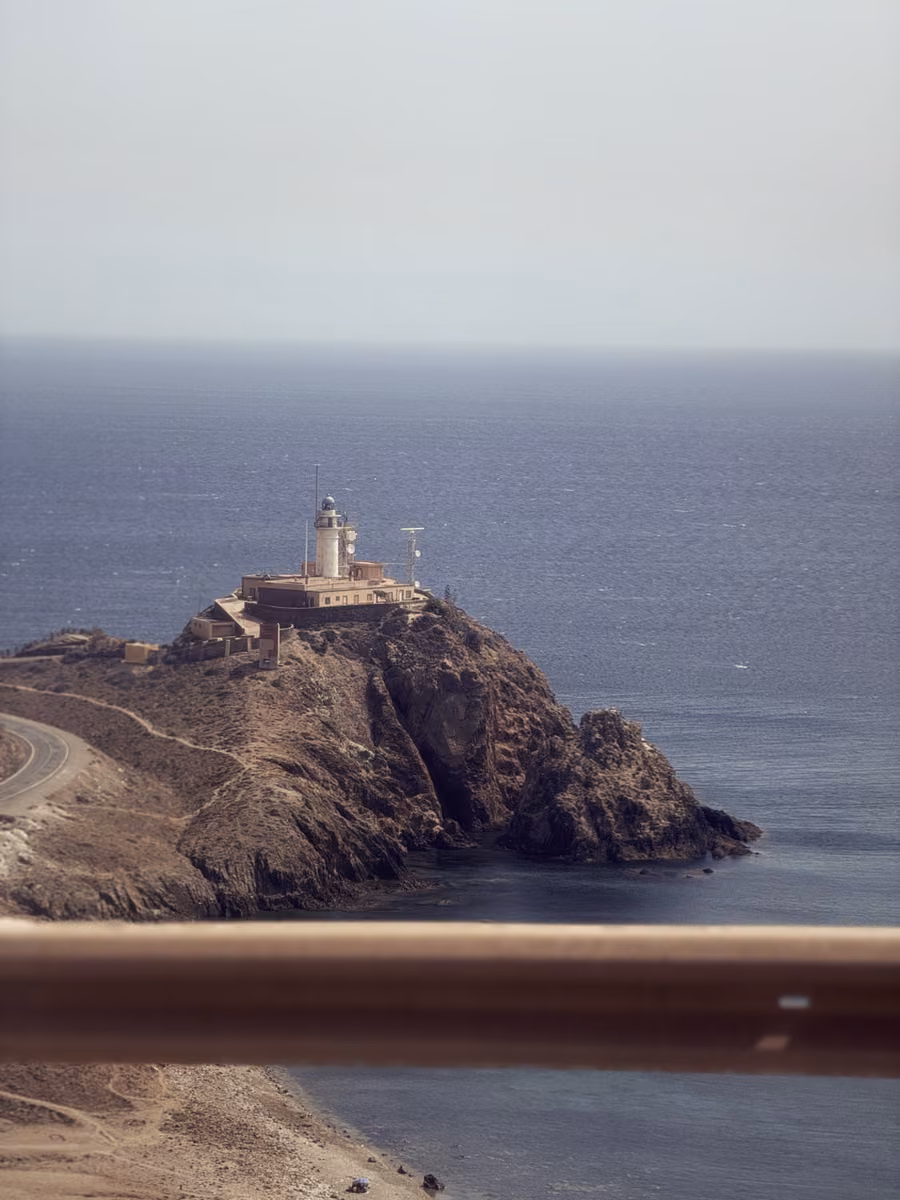 A white lighthouse on a rocky headland above the sea, seen over a road barrier.