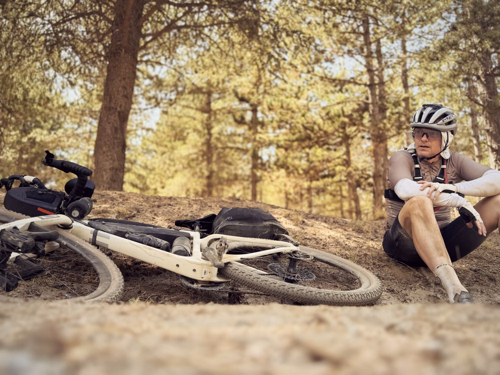 A rider sits in the shade of pine trees with his loaded bike lying beside him.
