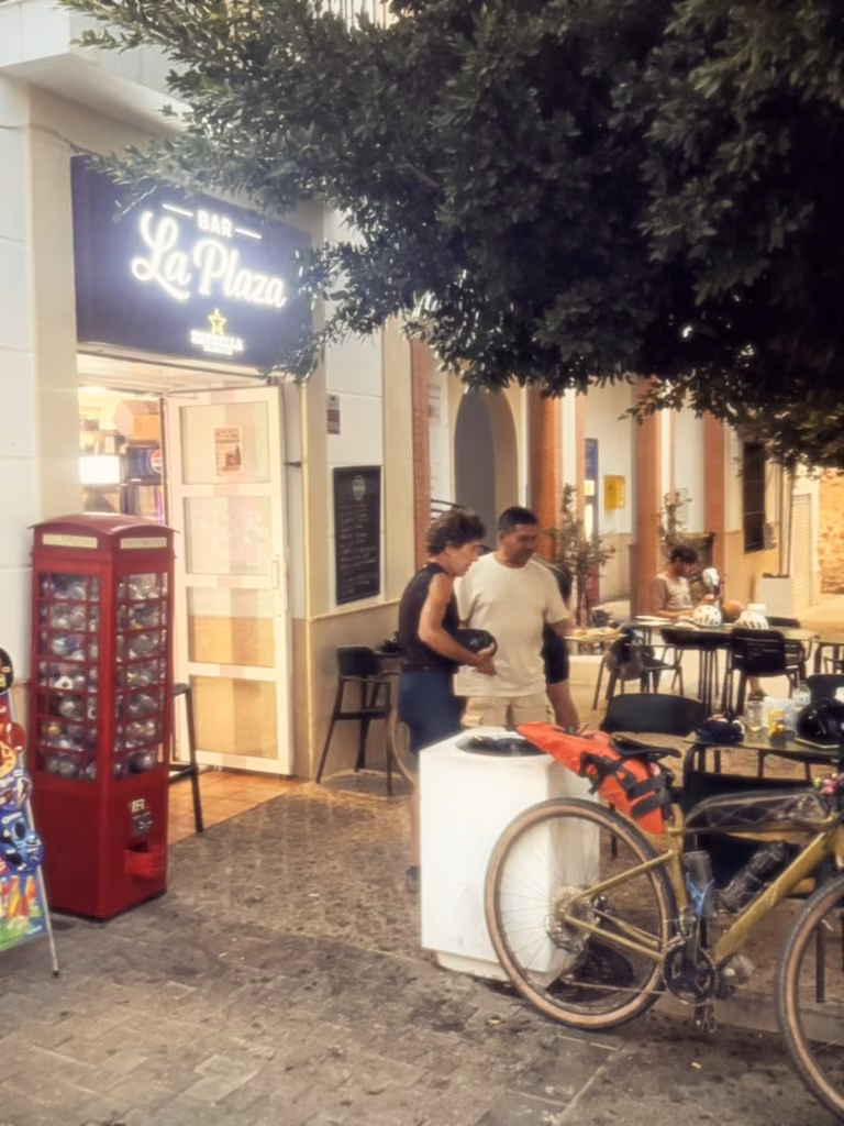 Bar La Plaza at dawn: riders at the tables under a tree, a loaded bike against a planter.