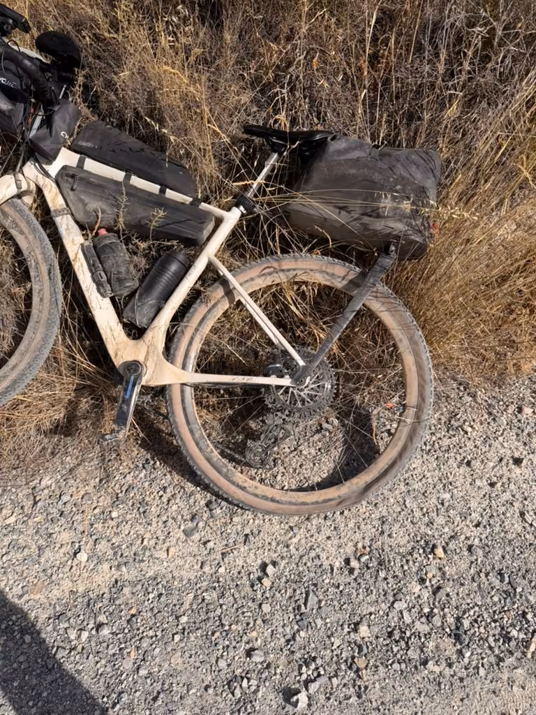 The loaded bike leans against dry grass beside a gravel track, its rear tire flat against the ground.