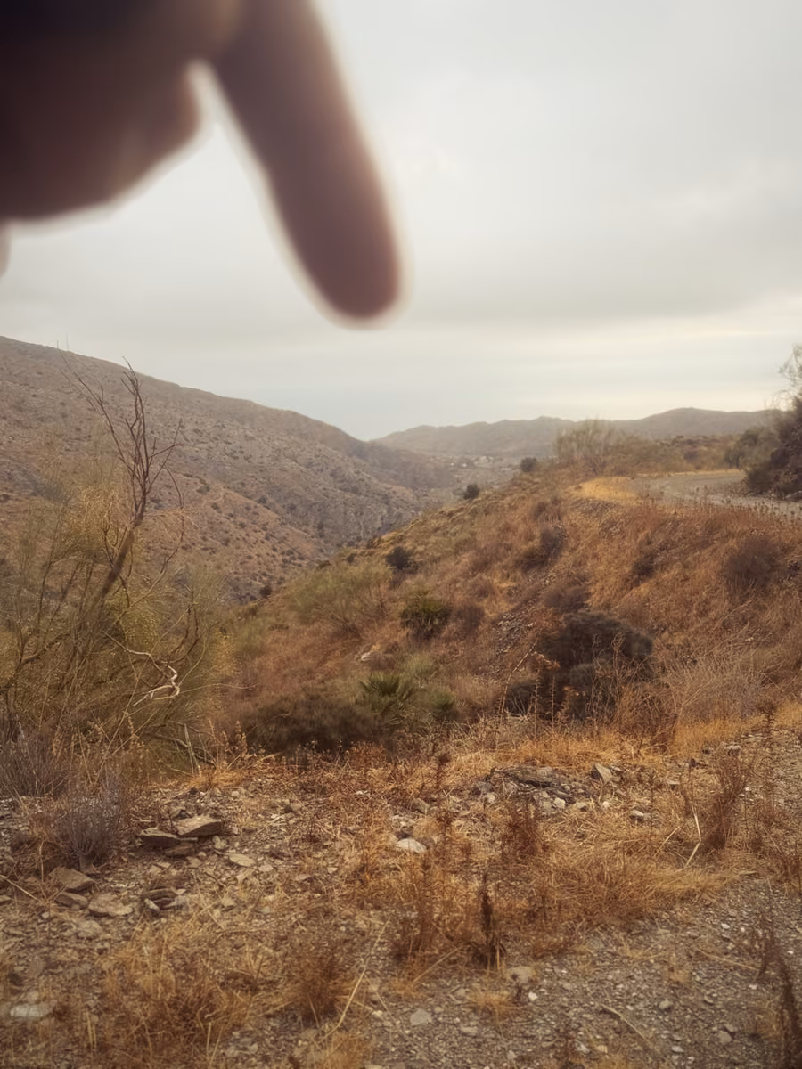 A finger pointing over a dry valley toward a faint gray strip of sea between the hills on the horizon.