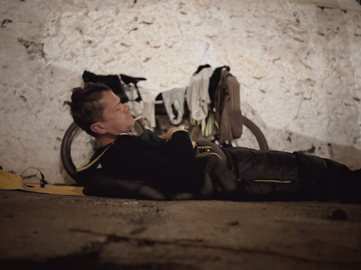 A rider lies in a sleeping bag on the ground against a whitewashed wall, with kit drying on the bike behind him and a flat yellow mat beside him.