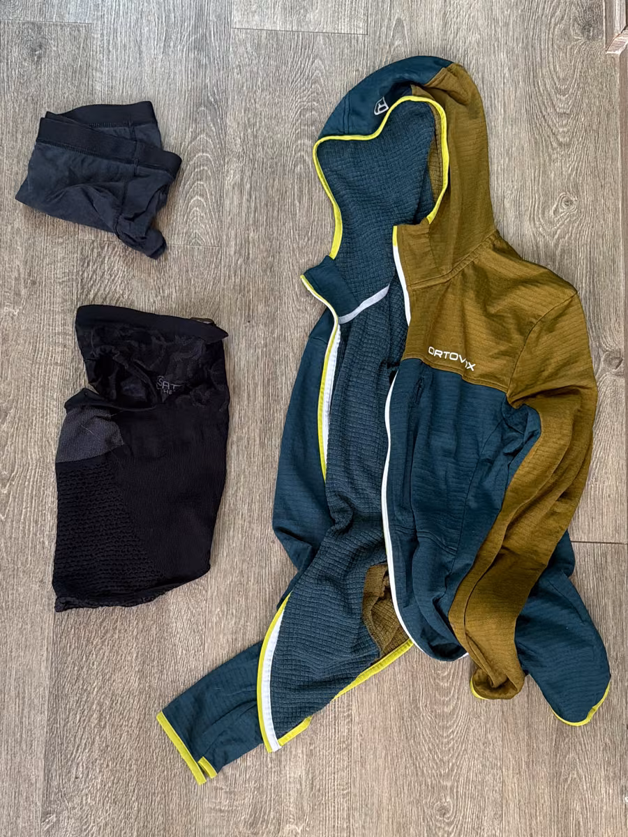A hooded layer and folded dark clothing laid out on a wood floor.