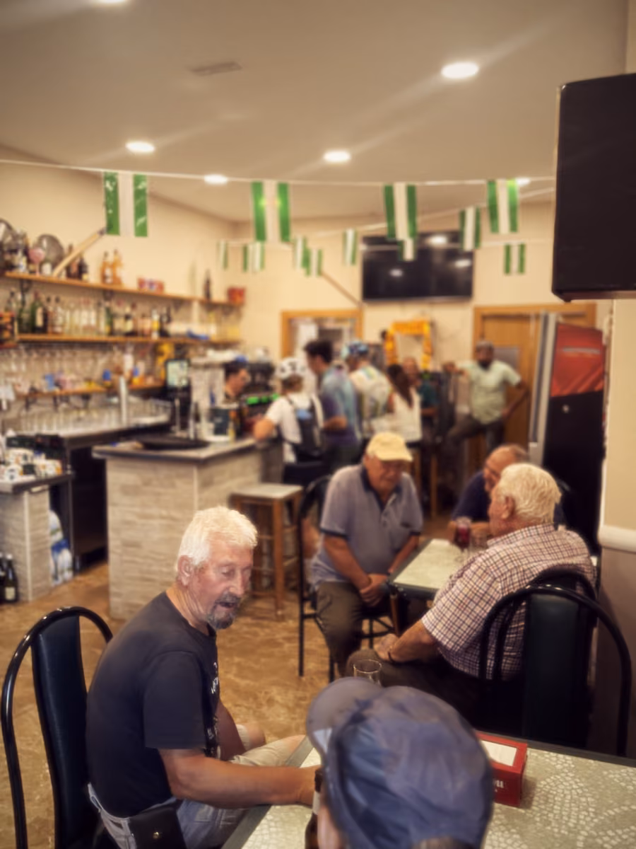 People sit at tables in a village bar while riders wait at the counter under green-and-white bunting.