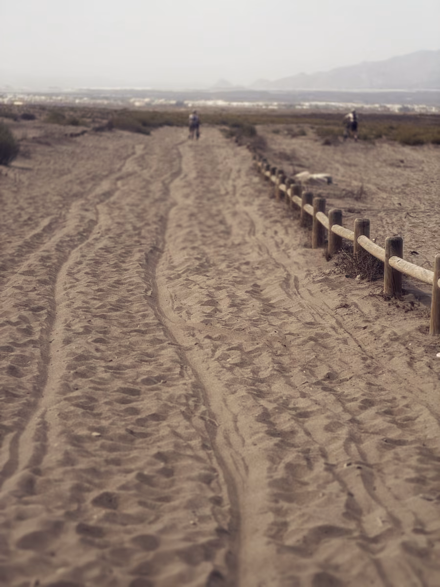 Deep tire tracks in a sandy path along a wooden fence, two riders far ahead, Almería's hills behind.