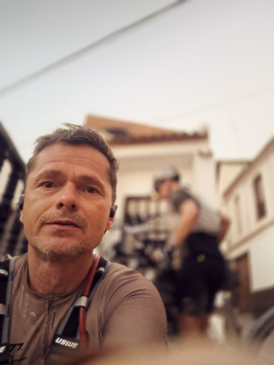 Close selfie of a rider with earbuds and a hydration vest, another rider packing his bike out of focus behind.