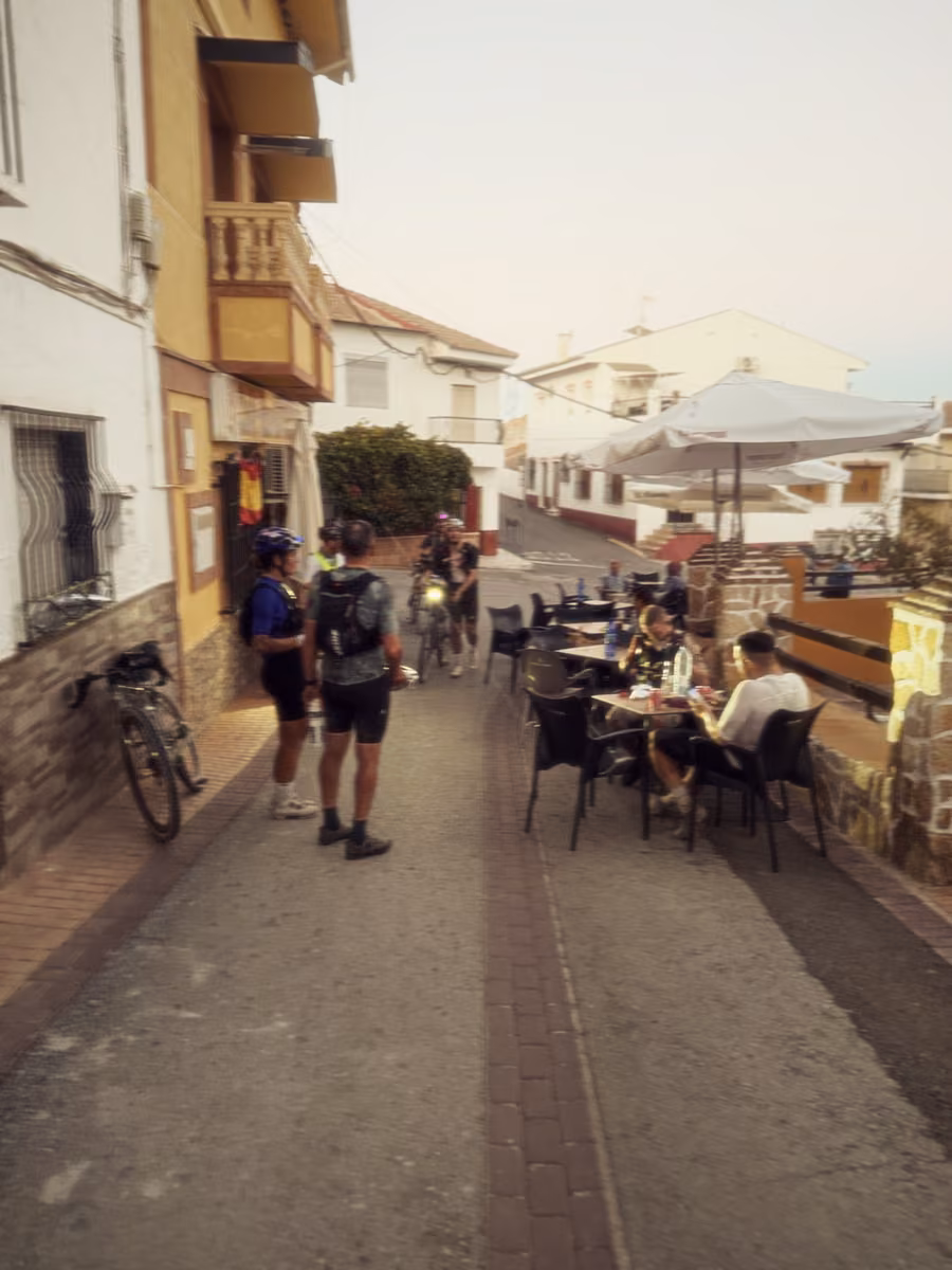 People gather outside a bar on a village street at dusk, with bikes leaning against the wall.